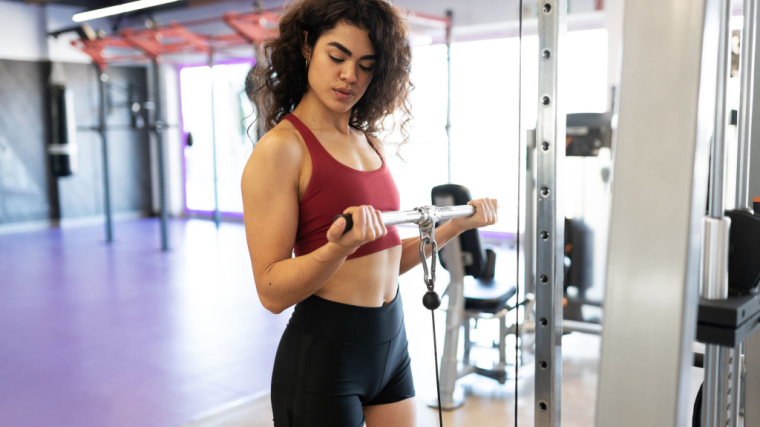 A person wearing a sports bra performs cable biceps curls in the gym.