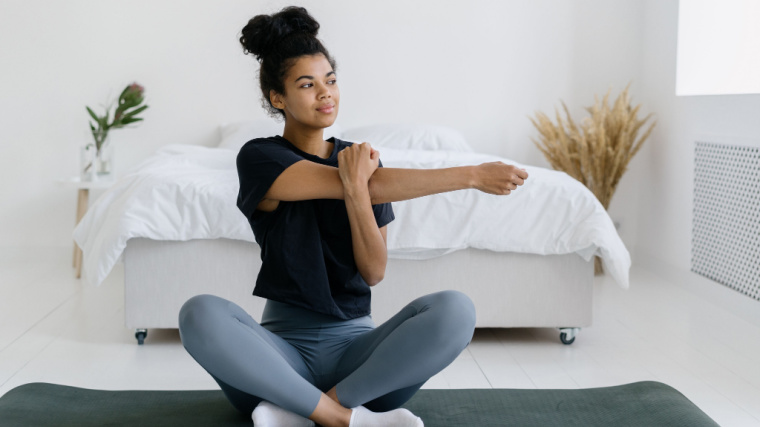 A person performs a seated triceps stretch on the floor in front of a bed.