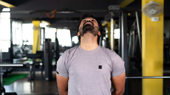 A person stands in the gym and stretches their neck by lifting their chin up and back.