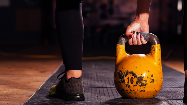 A close-up image shows a person with black and red nail polish gripping a yellow competition kettlebell.