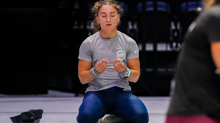Athlete, wearing a grey t-shirt and blue leggings, kneels in a meditative state.