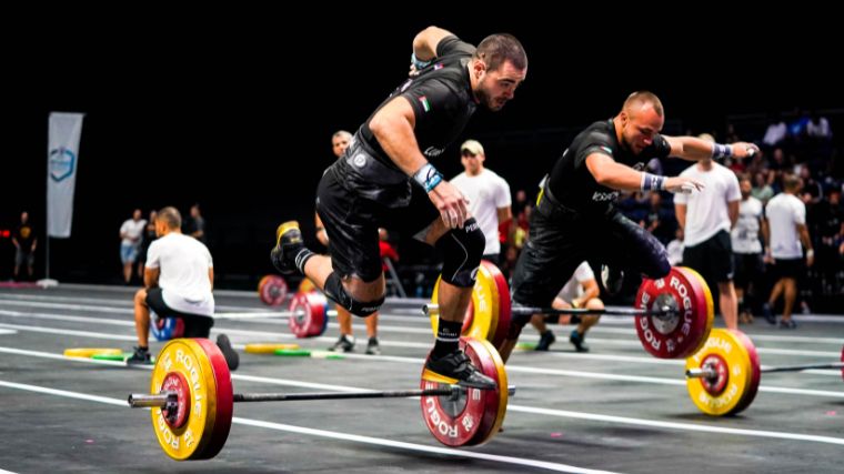 Two athletes leap over barbells loaded with red and yellow weight plates as they race to a finish line. 