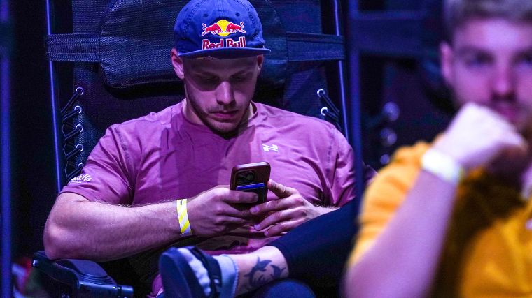 Man in purple t-shirt and a "Red Bull" hat sits on a chair and scrolls on his phone. 