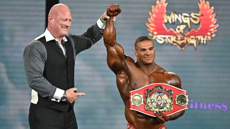 Bodybuilder has his hand raised while holding a red, gold-plated belt that signifies the "people's champion" at the 2022 Mr. Olympia competition.