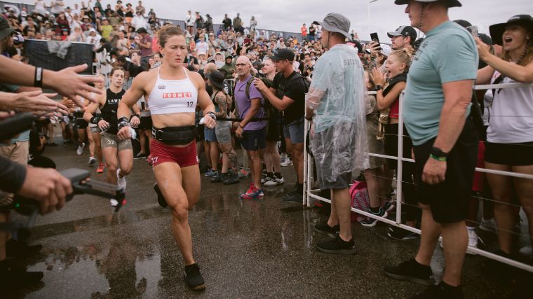 Tia-Clair Toomey running through a crowd of people with a focused look on her face, wearing a lifting belt, black sneakers, red shorts, and a white tank top.