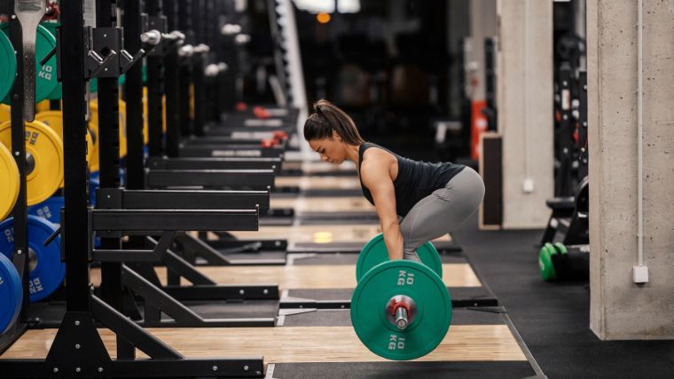 Sporty person wearing grey leggings and a black tank top doing Romanian Deadlifts 9the exercise) with a barbell loaded with one green 10-kilogram plate per side. 