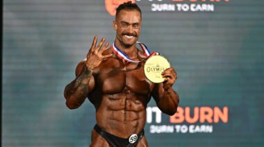 Bodybuilder Chris Bumstead holds his 2022 Classic Physique Olympia gold medal and four fingers to indicate his number of wins.