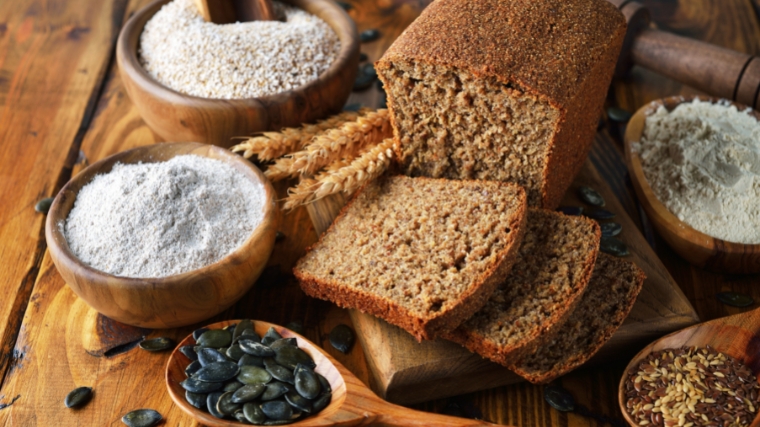 Partially sliced loaf of bread next to a few wheat stalks and bowls of flour