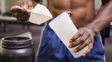 A shirtless person scoops pre-workout into a shaker bottle.