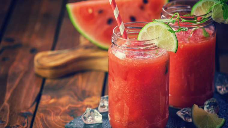 A jar full of watermelon juice sits on a table with a watermelon and cutting board.