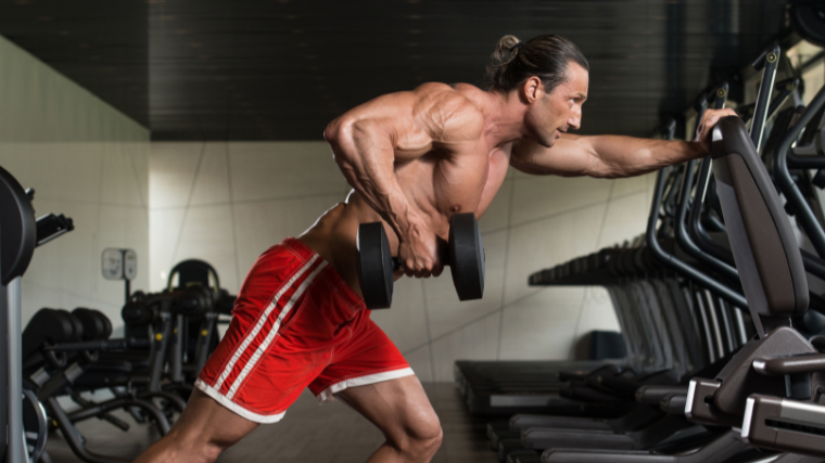 Muscular shirtless man performs dumbbell row while leaning on cardio equipment