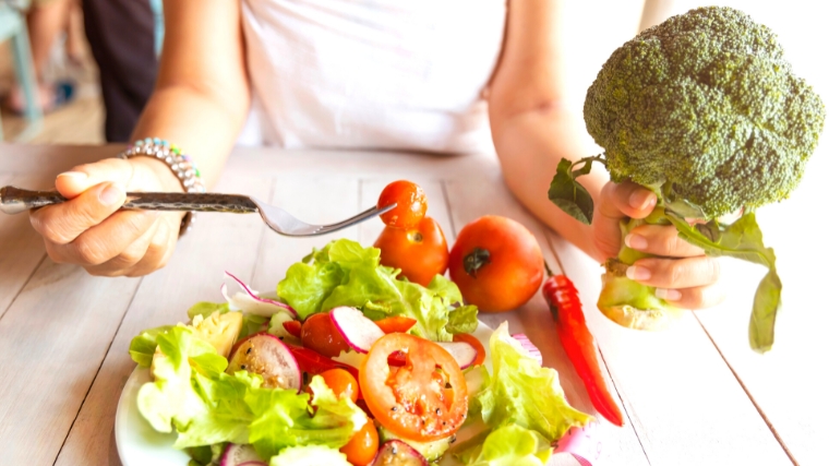 A person using a fork on a salad, and holding a broccoli on the other hand.