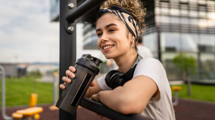 Person smiling leaning on a squat rack with shaker bottle in hand and headphones around neck
