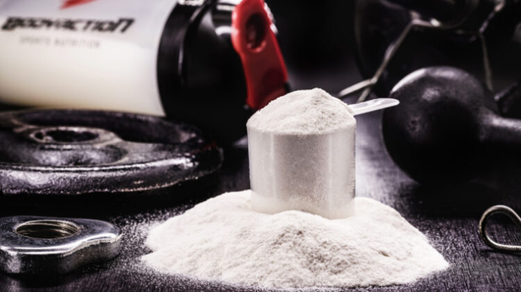 Scoop of creatine in a scooper with powder around it in front of a shaker bottle and kettlebell