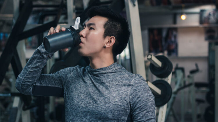 Person drinking from shaker bottle in the gym with weight machines behind them in a gray tight hoodie