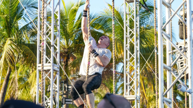 An athlete performs a rope climb in front of palm trees.
