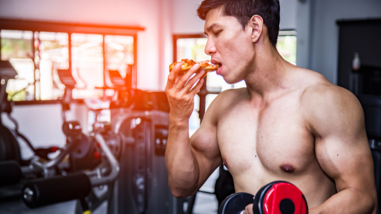 A shirtless person eats pizza in the gym.