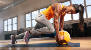 A woman in an orange shirt performing an exercise with a medicine ball in the gym.