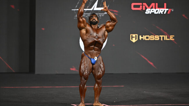 2022 Mr. Olympia Hadi Choopan posing on stage in Las Vegas with his arms extended overhead.