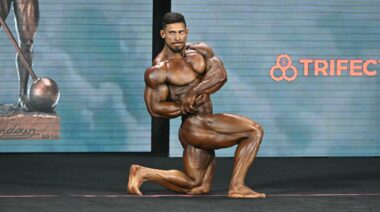 Muscular bodybuilder kneeling on one leg while performing a side chest pose on stage.
