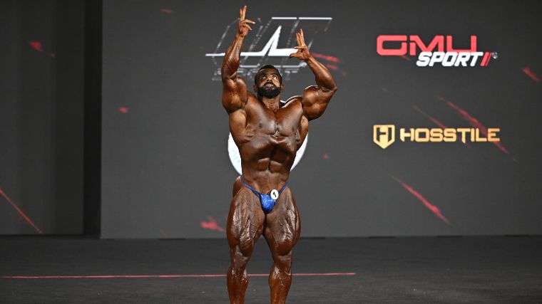 Muscular bodybuilder posing on stage with his arms extended over their head.