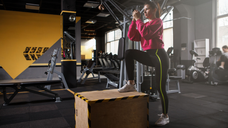 Person in pink sweater and leggings steps up onto plyo box with hands clasped in frnt of their chest
