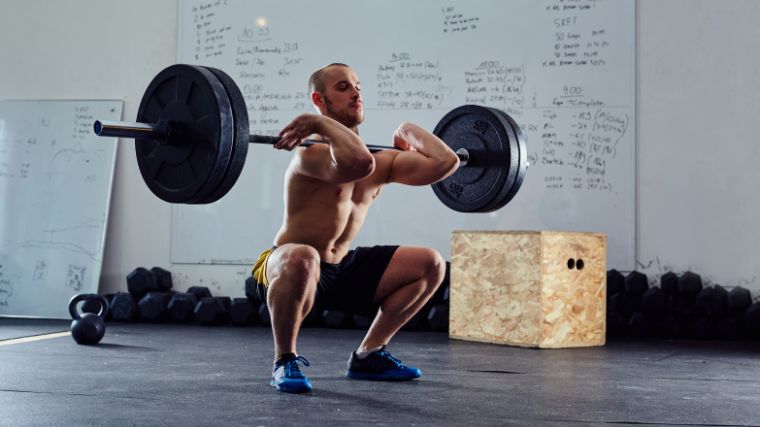 Shirtless person in yellow and black shorts performs a front squat with a loaded barbell.