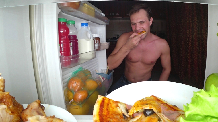 A shirtless person binge eating at an open fridge.