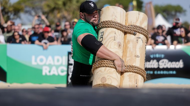 Strongman in a green shirt and elbow sleeves carries a bundle of logs to load onto a platform.