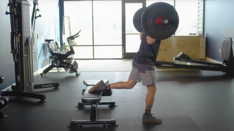 A person starting to do a barbell Bulgarian split squat.