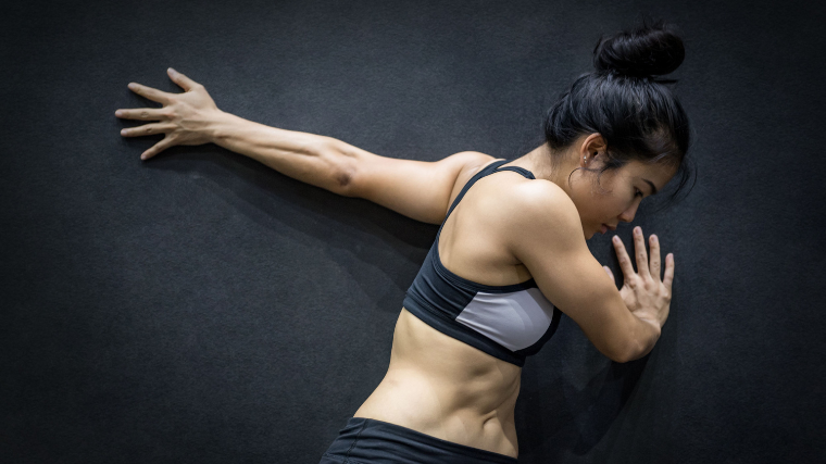 An athlete wearing a sports bra performs a biceps stretch on a wall.
