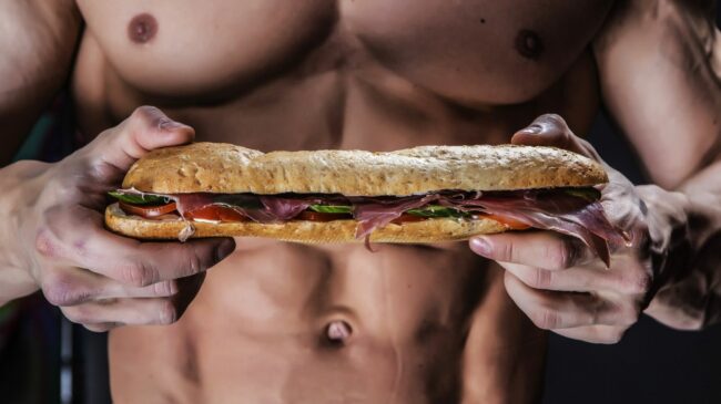 A bodybuilder holding an Italian-style sandwich in front of their shirtless torso.