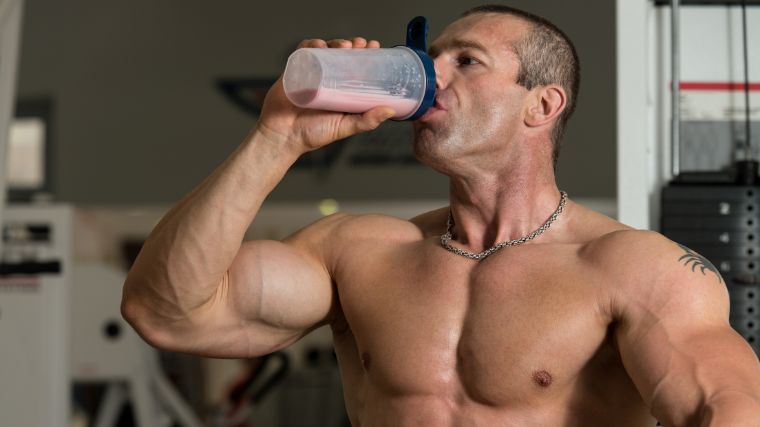 A shirtless bodybuilder drinking whey protein supplement from a shaker bottle.