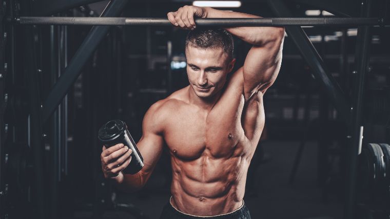 A bodybuilder holding a shaker bottle while leaning on a bar.