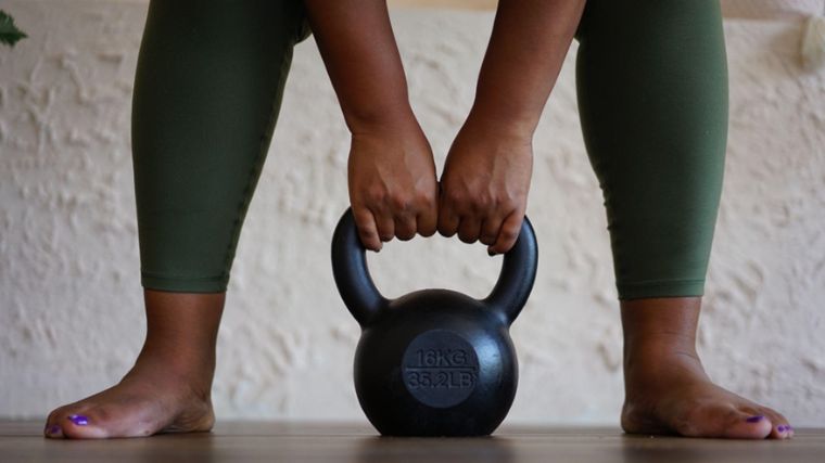 A close up of hands grabbing a kettlebell on the floor between feet that are hip-width apart.