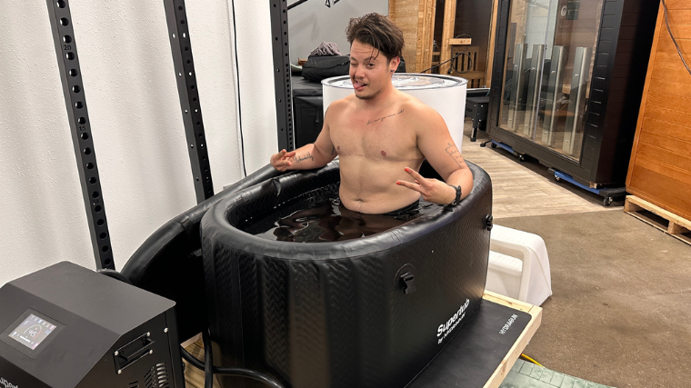 A shirtless person with top surgery scars sits in the Hydragun Supertub.