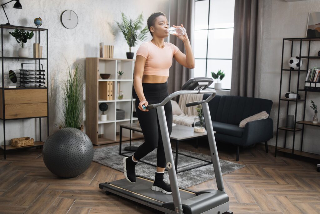 A woman in workout attire walking on a treadmill in her home while drinking water.