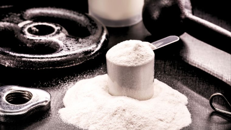 Whey protein powder in a scoop, set beside weights and spilled whey powder.