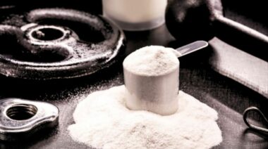 Whey protein powder in a scoop, set beside weights and spilled whey powder.
