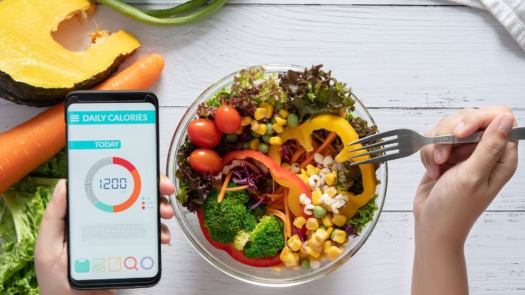 Fresh fruits and vegetables in a bowl and a hand holding a smartphone showing a food calorie tracker.