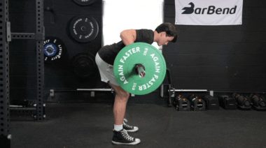A person wearing grey shorts and a black t-shirt perform bent-over barbell rows with a green, 25-pound plate on each end.