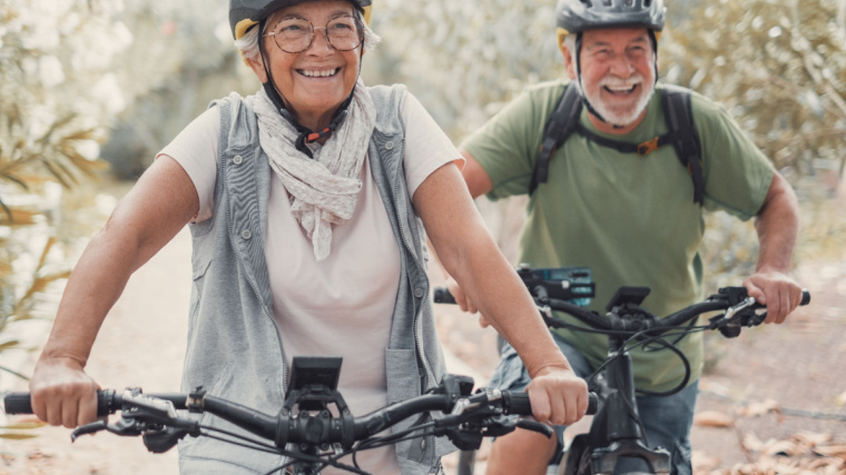 Two elderly people wearing helmets and happily riding bicycles.