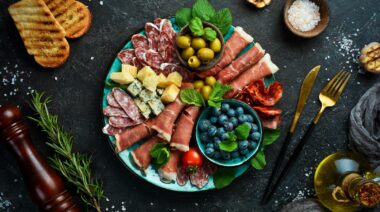 Plate with cheese and ham, prosciutto, jamon salami, and snacks. On a black stone background.