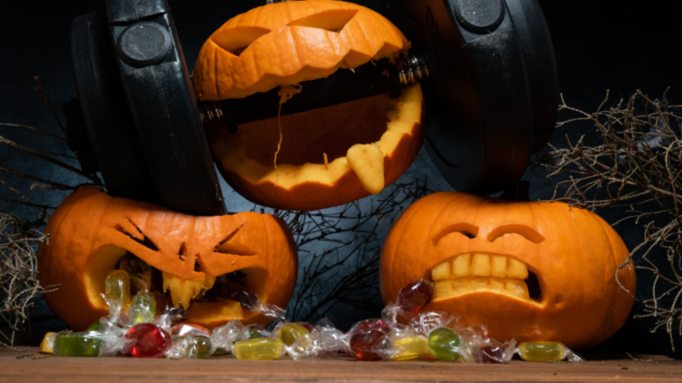Halloween pumpkin clenching teeth on dumbbell, crushing other carved Jack-o'-lanterns with candies falling out of its mouth.