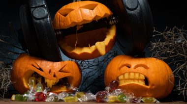 Halloween pumpkin clenching teeth on dumbbell, crushing other carved Jack-o'-lanterns with candies falling out of its mouth.