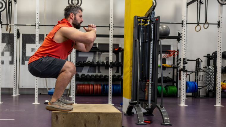 A bearded person jumping up on wooden box in a CrossFit gym.
