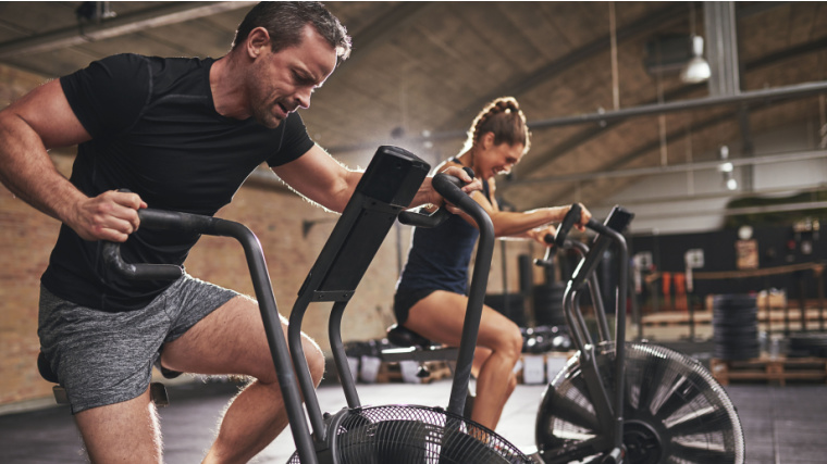 Two people training hardly on cycling machines in light spacious gym.