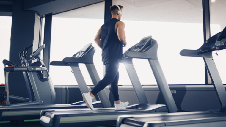 A profile view of a person listening to music with headphones while jogging on a treadmill in amodern gym.