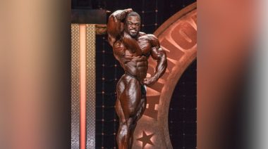 2019 Mr. Olympia winner Brandon Curry performs an abdominal and thighs pose on stage, wearing posing trunks.