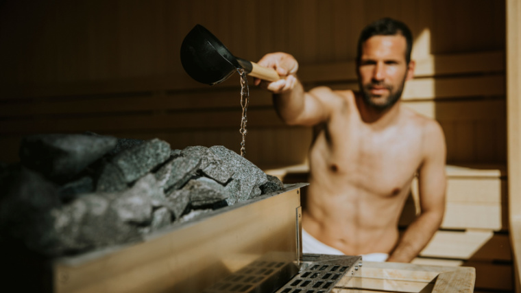 A fit person pouring water unto hot stone in sauna. 